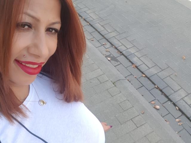 IssaDiamondd with red lipstick, nose piercing, white shirt, and shoulder bag, smiling at the camera, showing her tits slightly, on a grey brick sidewalk with leaves.