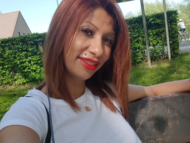IssaDiamondd met rode lippenstift, een neusring, een wit shirt en bruin haar, lachend, leunend tegen een donker houten oppervlak, waarbij haar kleine borsten zichtbaar zijn, in een zonnige buitenomgeving..