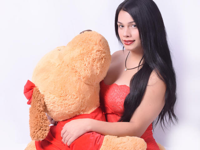 ValeriaHot69TS met lang zwart haar, een rode kantjurk en een ketting, die een grote, oranje, pluizige teddybeer met een rode strik omhelst, en die lichtjes glimlacht. Haar borsten zijn zichtbaar..