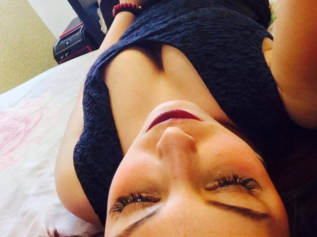 MissAnalis, avec son visage mignon, son rouge à lèvres rouge foncé et ses longs cils, est allongée sur un lit, vêtue d'un haut bleu foncé sans manches, qui révèle ses petites seins fermes, et elle porte un bracelet rouge..