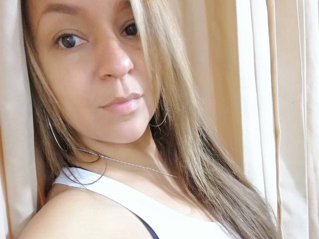 Angelonix with light brown hair, small nose, and silver hoop earrings, wearing a white tank top, has a subtle smile and is looking at the camera.