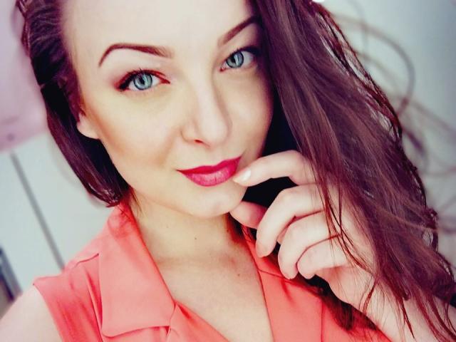 StellaLee hat blaue Augen, roten Lippenstift und braunes Haar, trägt ein pinkes Hemd und hat den Finger am Kinn, während sie mit einem leichten Lächeln in die Kamera schaut..