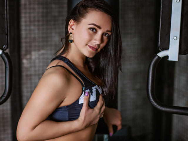 Chantal, de cabello oscuro, sosteniendo sus pequeños y firmes senos con un sujetador deportivo oscuro, sonriendo en un gimnasio, con aretes verdes, con una mirada sutil y sexy..