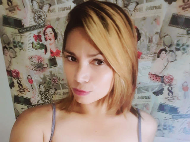 NatashaRomanof with shoulder-length blonde hair, pink lipstick, and brown eyes, wearing a gray spaghetti strap top, showing a slight smile, looking at the camera.