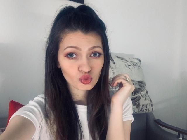 BlossomPussy with pouty pink lips, long dark brown hair in a high ponytail, wearing a white tee, holding a ring on her right hand, looking at the camera.
