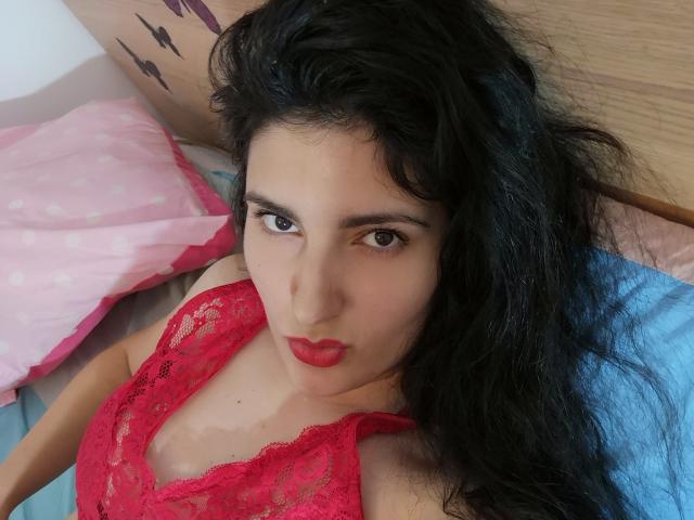 AddictedToUTS, avec de longs cheveux noirs, un haut en dentelle rouge et du rouge à lèvres, est allongée sur un lit avec des oreillers roses et bleus. Ses seins sont visibles, ses lèvres sont légèrement entrouvertes, elle a les yeux marron et une expression sérieuse..
