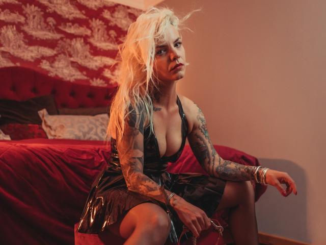 VanessaOdette, avec ses cheveux blonds et en désordre, ses tatouages et sa robe en latex noir, assise sur un lit rouge, regardant sur le côté, montrant sa poitrine, avec des bracelets à son poignet droit..