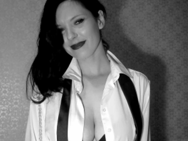 Lucy Violet, avec du rouge à lèvres foncé, à moitié souriante, portant une chemise déboutonnée qui révèle ses grandes seins, des bretelles noires, et de longs cheveux noirs, sur une photo en noir et blanc..