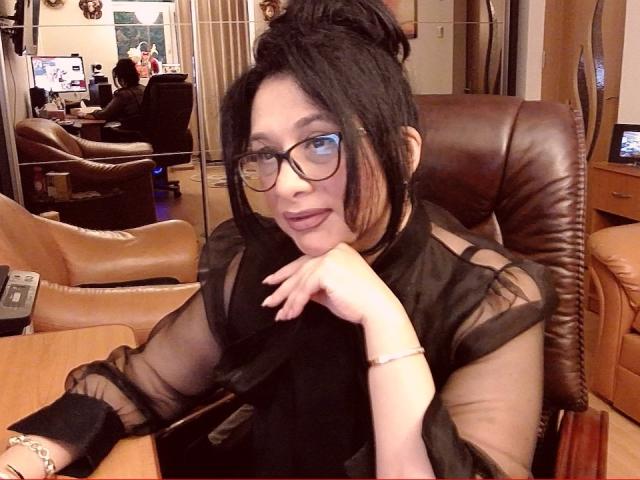 ClassybutNaughty with black hair in a bun, glasses, and a sheer black top, resting chin on hand, tongue slightly out, sitting in a brown leather chair.