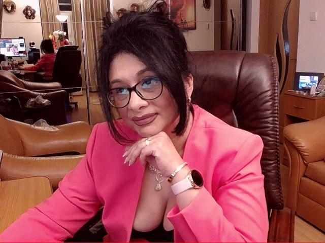 ClassybutNaughty with black hair, glasses, pink blazer, and cleavage in a brown leather chair, resting her chin on her hand, wearing a pink watch, and a ring.