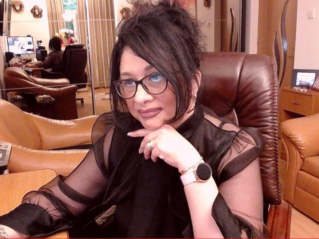 ClassybutNaughty, met zwart haar, een bril en een doorschijnend zwart topje, leunt ze haar kin op haar hand, waarbij haar roze lippenstift en een polshorloge zichtbaar zijn, terwijl ze in een leren stoel zit..