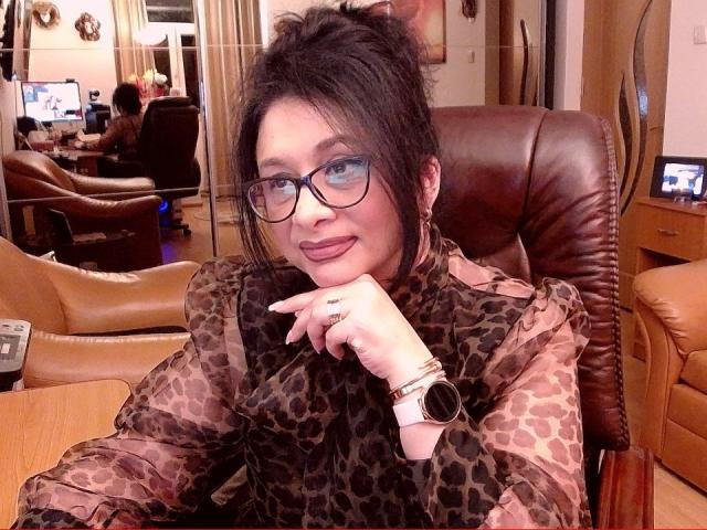 ClassybutNaughty with glasses, dark hair in a messy bun, leopard print shirt, pink lipstick, finger on chin, ring, bracelet, and watch, smiling in a brown leather chair.
