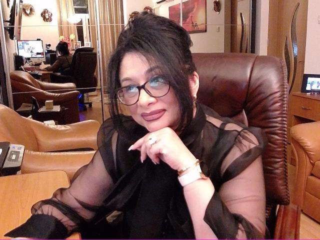 ClassybutNaughty with dark hair, glasses, and deep red lipstick, wearing a sheer black top, sits in a brown leather chair, hand on chin, looking cute and sexy.