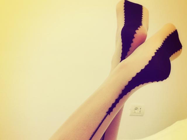 MistressOfShadow's legs, crossed at the ankles, wearing sheer black stockings with a zigzag pattern, highlighting her pale skin. Her feet are pointed upwards, showing off her sexy legs.