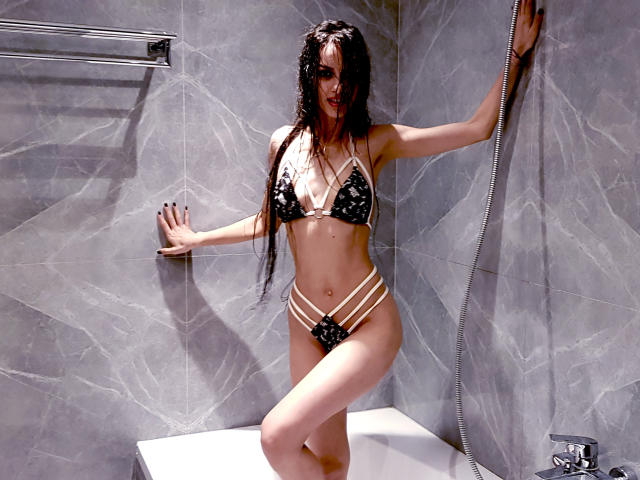 KatherineBisou with wet black hair, wearing a black and gold strappy bikini, standing in a gray marble shower, one hand on the wall, the other on her ass.