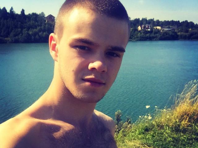 MarcusFate, torse nu, avec des cheveux bruns courts, une peau claire et une légère barbe de quelques jours, se tient près d'un lac bleu, regardant légèrement sur le côté avec une expression neutre..