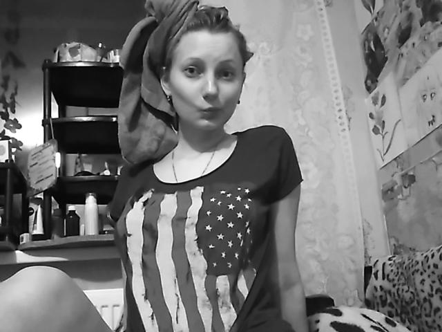 JillFox avec une serviette sur la tête, portant une chemise sombre avec un imprimé du drapeau américain, assise dans une pièce en désordre, photo en noir et blanc, petit nez, écouteurs dans les oreilles, expression boudeuse..