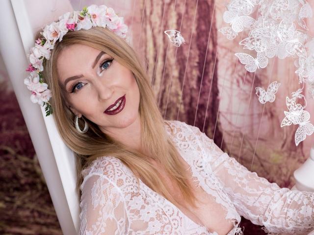 LadyLea with blonde hair, flower crown, and hoop earrings, wearing a white lace top, smiling with dark red lipstick, showing her tits through the lace.