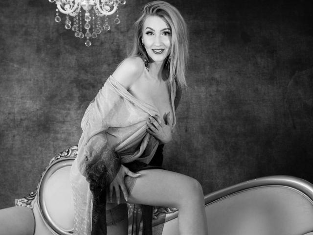 LadyLea, with medium tits, sits on an ornate chair, wearing a sheer robe, exposing her right boob, smiling, legs apart, chandelier above, black-and-white photo.