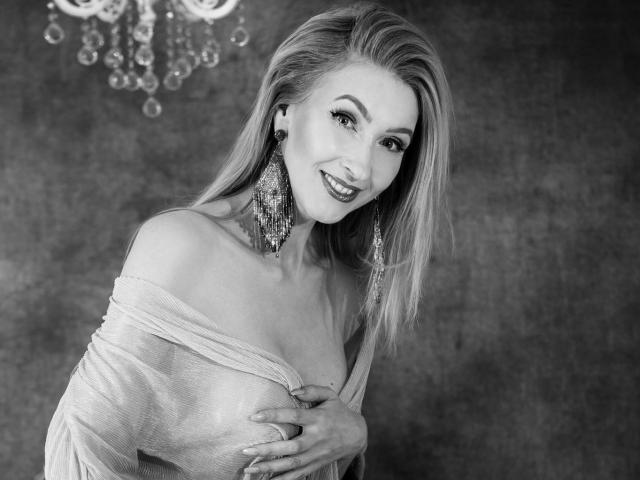Black and white photo of LadyLea with long blonde hair, smiling, wearing off-shoulder top, large chandelier earrings, hand covering her tits, sexy, elegant, and confident.