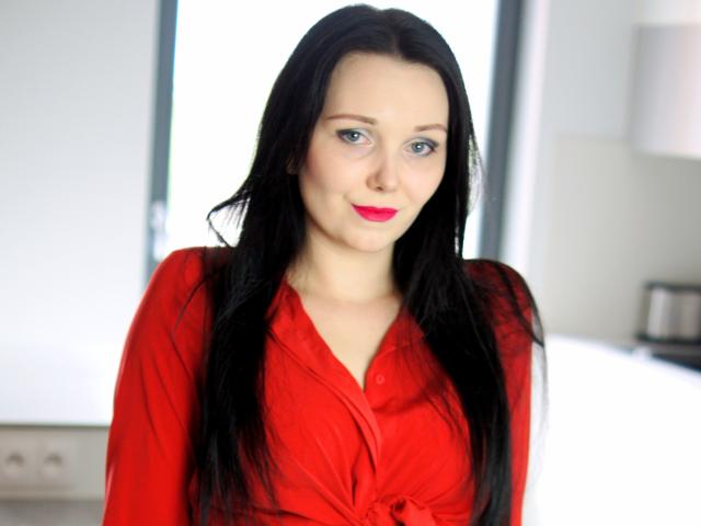 QueenZoe with long black hair, red lipstick, and a red button-up shirt, smiling softly at the camera, showing her tits subtly tied in the shirt.