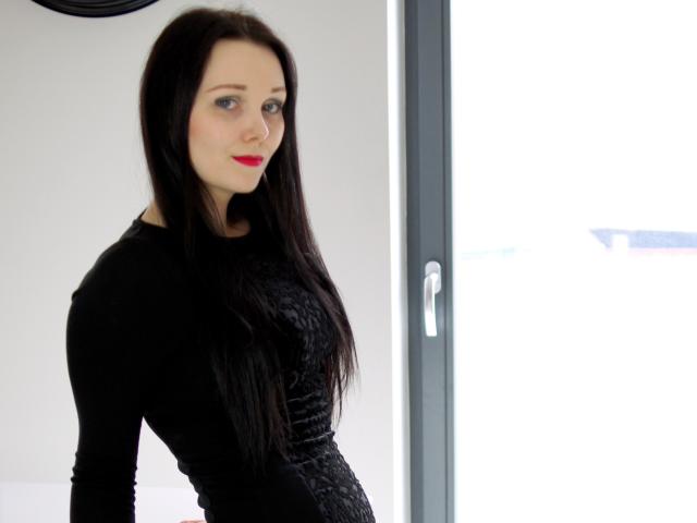 QueenZoe, avec ses longs cheveux noirs, portant une robe noire moulante, du rouge à lèvres et des yeux bleus, se tient près d'une fenêtre lumineuse. Ses fesses sont légèrement visibles sous la robe..
