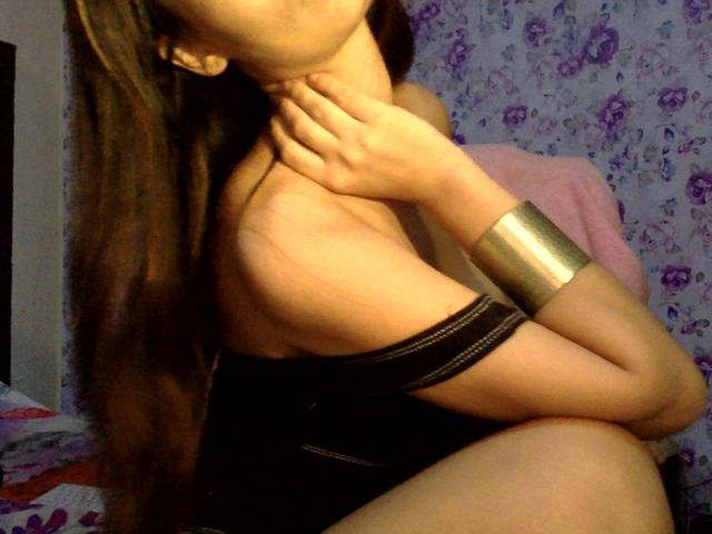 GiovannaSweet, com longos cabelos castanhos, tocando o pescoço, vestindo um vestido preto com alças e um bracelete dourado brilhante, mostrando o ombro nu e parte do seio..