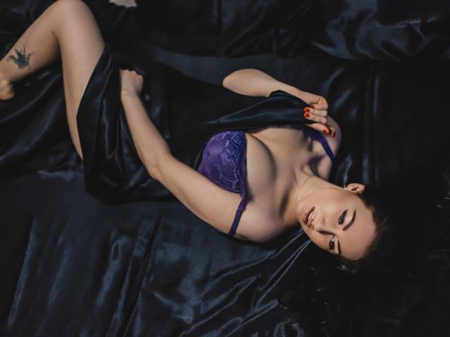 TellySabe lying on black silk, wearing purple lace bra, holding black fabric over her tits, red nail polish, tattoo on left thigh, dark hair, sultry expression, dark lighting.