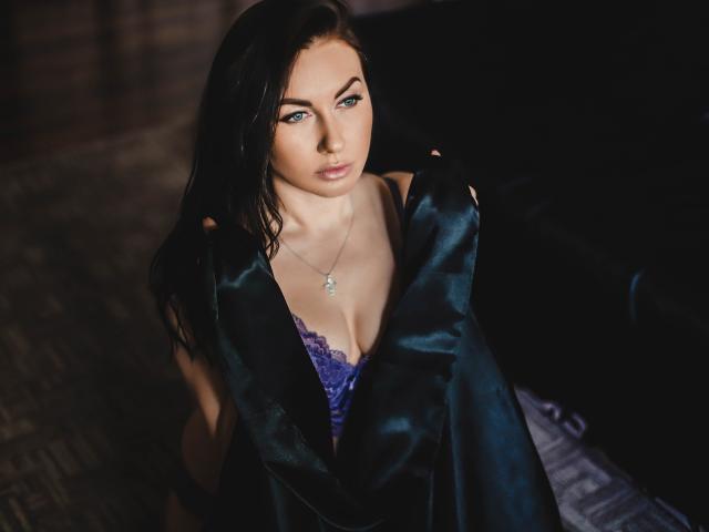 TellySabe, con i capelli neri e umidi, gli occhi azzurri e un reggiseno di pizzo viola, avvolta in una veste di raso nero, che indossa una collana d'argento, seduta su una superficie scura e strutturata..