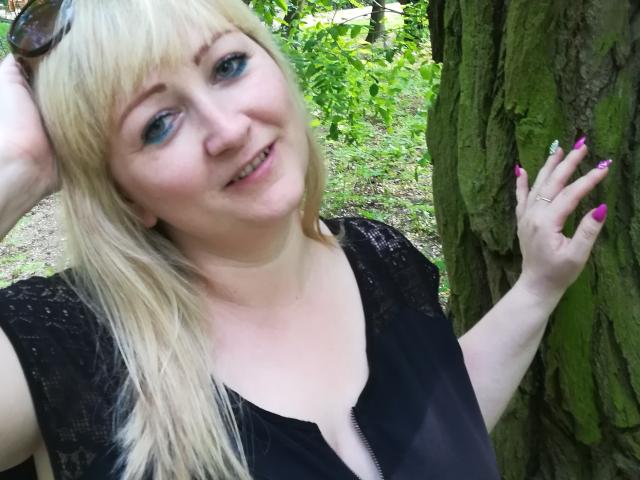 MarinaSweet, con cabello rubio, ojos azules, blusa de encaje negro, uñas rosas, anillo, sonriendo, mano en un árbol, gafas de sol en la cabeza, fondo de bosque, mostrando ligeramente los senos en la blusa escotada..