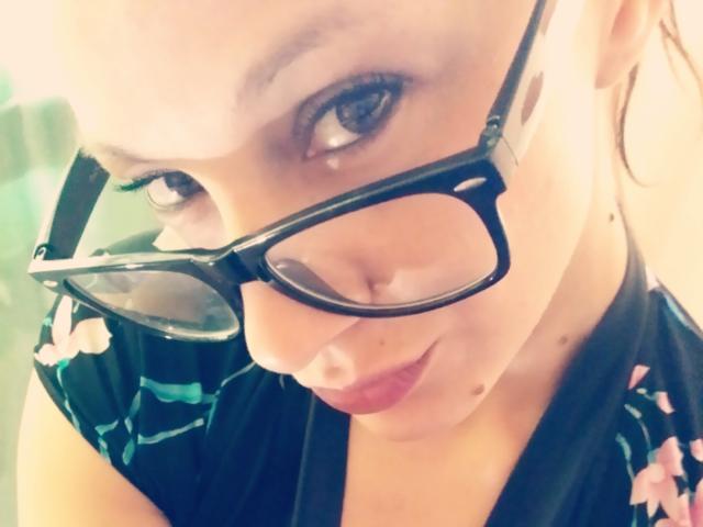 CassandraMichelli with black glasses, looking up, wearing a dark floral dress, smiling slightly, her nose and lips close to the camera, showing a cute, nerdy, sexy look.