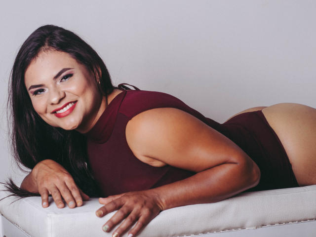 Briana Preston est allongée sur un banc blanc, souriant avec du rouge à lèvres, vêtue d'une combinaison rouge foncé, ses fesses et ses jambes étant visibles, avec des cheveux noirs, une peau brune et une pose détendue..
