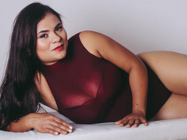Briana Preston è sdraiata su una superficie bianca, indossa una tuta trasparente color borgogna che mette in risalto il seno e i glutei, con lunghi capelli scuri e rossetto rosso..