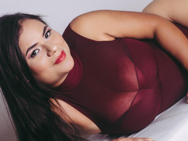 Briana Preston, con i capelli scuri, il rossetto rosso e una maglia marrone trasparente, è sdraiata su una superficie bianca, mostrando il seno e un sorriso dolce. Ha un viso carino..