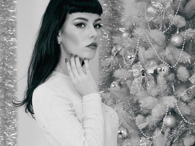 AmberBrowns with dark, straight hair and blunt bangs, wearing a white cardigan, standing by a decorated Christmas tree, looking at the camera with her hand near her face.