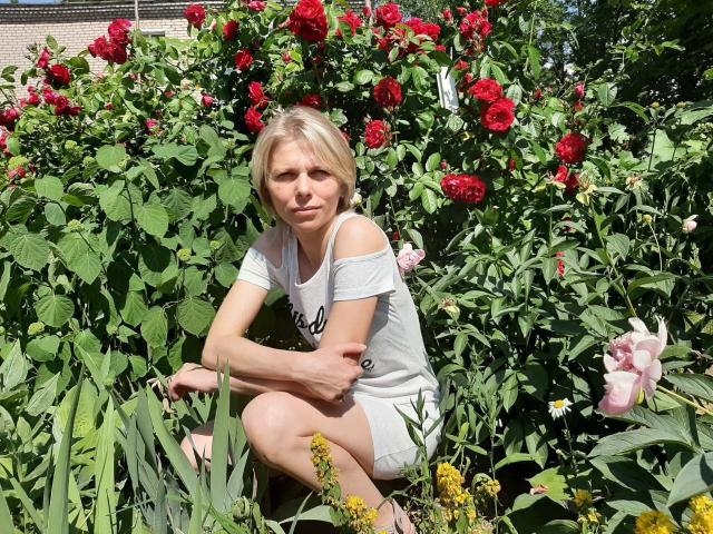 VandaFlower has blonde hair, small tits, and is crouching in a garden with red roses, wearing a white off-shoulder top and shorts, surrounded by greenery and yellow flowers.