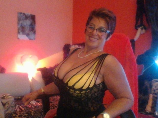 Bettina, a curvy woman with big tits, wearing a black, strappy, lace-up dress, glasses, and a necklace, smiles in a red-lit room with a red chair.