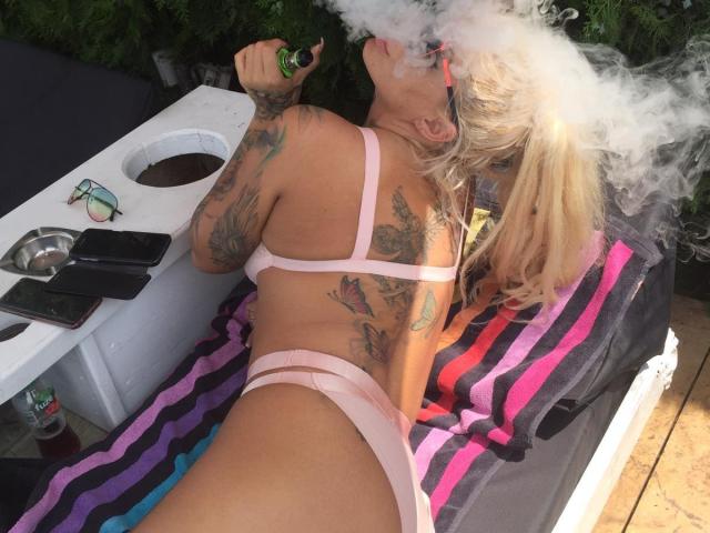 BarbaraBae con i capelli biondi, biancheria intima rosa con spalline sottili, tatuaggi, che fuma uno spinello, sdraiata su un asciugamano a righe, tavolo all'aperto con bevande e telefono, con i glutei sollevati, pavimento in legno..