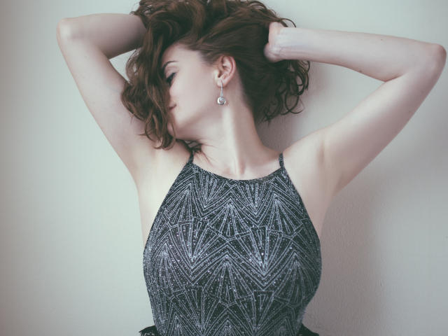 ChicEva con los brazos levantados, cabello castaño ondulado, vistiendo un top negro con lentejuelas de tirantes, que muestra sus senos y sus delgados brazos, con aretes colgantes, en una luz suave..