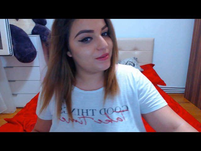 Adeline, con i capelli castano chiaro, una maglietta bianca e un letto rosso, ha un sorriso carino, un seno piccolo e una corporatura leggermente robusta, ed è seduta in una camera da letto con un peluche..