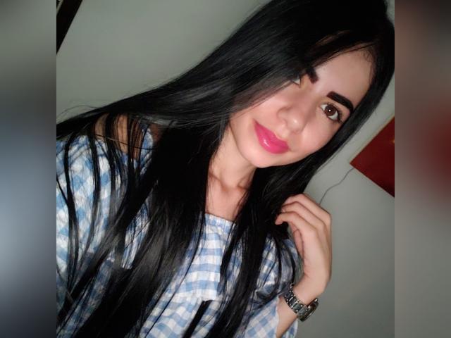 LexyJensen with long black hair, blue checkered shirt, pink lipstick, brown eyes, and a silver watch, smiling slightly, touching her hair with one hand.