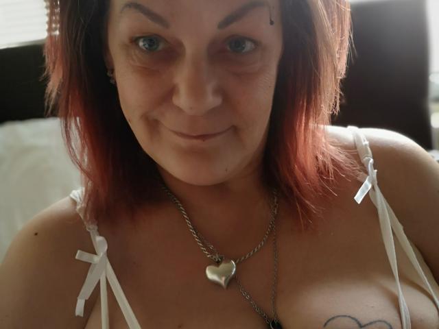 LatexShantie met rood geverfd haar, blauwe ogen en een klein beauty mark, draagt een witte top met bandjes, een hartvormige ketting en heeft een zichtbare tatoeage op haar borsten..