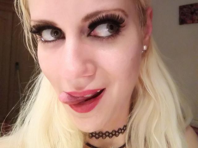 SwissMiss hat blondes Haar, große blaue Augen mit dicken schwarzen Wimpern und steckt ihre Zunge heraus, wobei sie roten Lippenstift trägt. Sie trägt eine schwarze Halskette und Diamant-Ohrringe..