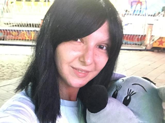 AmethystCharm with black hair, light skin, smiling, holding a gray cat-stuffed toy with black ears, wearing a light blue shirt, in a bright outdoor setting.
