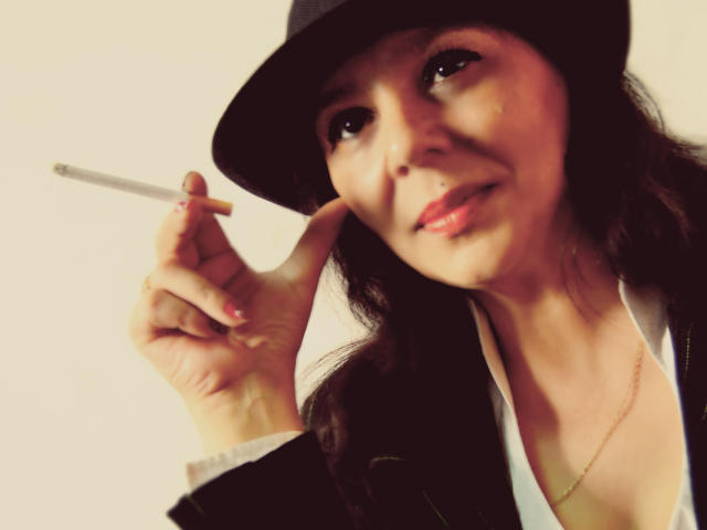 SxyVivian with dark hair, wearing a black hat, holding a cigarette in her right hand, slightly smiling, wearing a black jacket and white shirt, necklace visible.