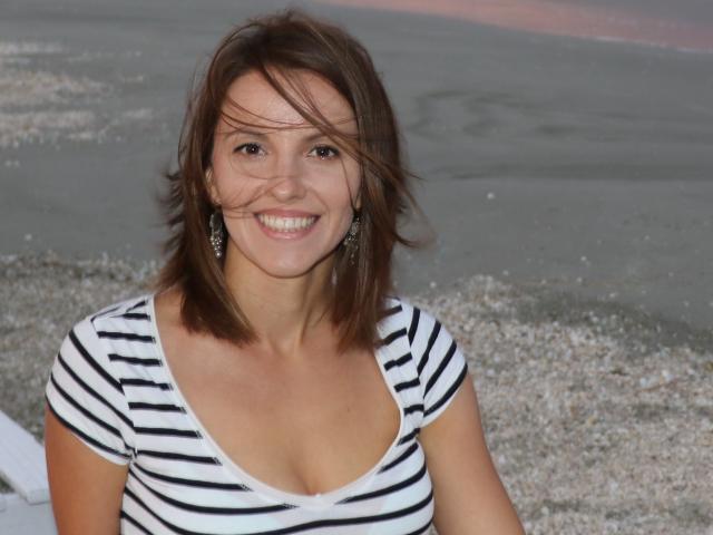 MissJoliSourireBg heeft bruin haar, ze glimlacht, draagt een gestreepte top in wit en zwart, waarbij haar kleine borsten zichtbaar zijn, ze draagt oorbellen en staat op een kiezelstrand..