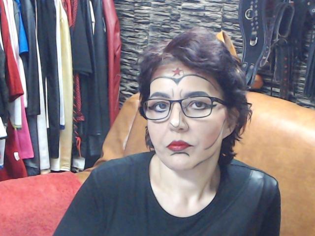 SexyLilly69 with short brown hair, glasses, red lipstick, black shirt, nose ring, and makeup with a red star on her forehead, sitting on a brown leather seat.