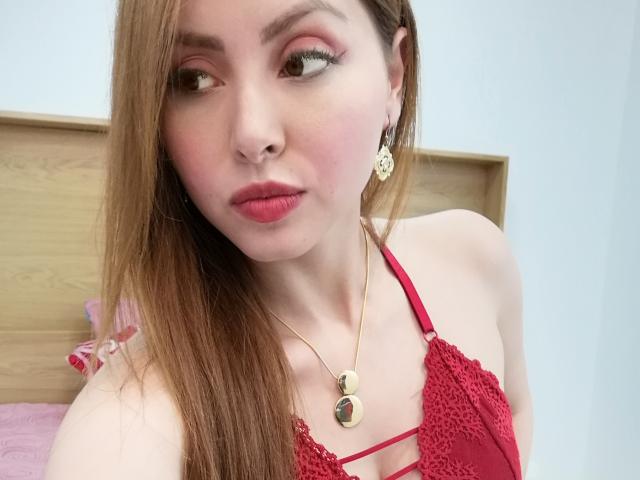 Katerina Salvatore met lang bruin haar, roze oogschaduw, rode lipstick, een rode kanten bh, een gouden ketting en bloemenoorringen, kijkt opzij, met volle lippen, bleke huid..