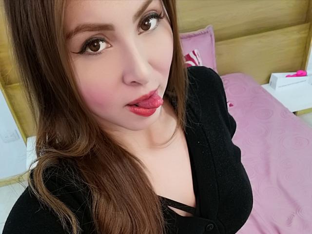 KaterinaSalvatore with brown hair, sticking out her red tongue, wearing a black shirt, showing her large boobs, and pink blush on her face, in a bedroom.