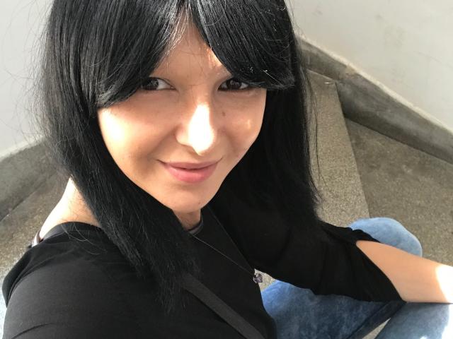 AmethystCharm with black hair, smiling at the camera, wearing a black shirt and blue jeans, sitting on concrete steps, her cute face and small nose in the shot.